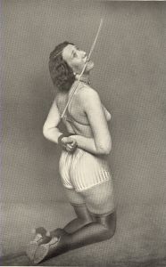 Kneeling submissive woman with hands tied, holding a cane in her mouth.